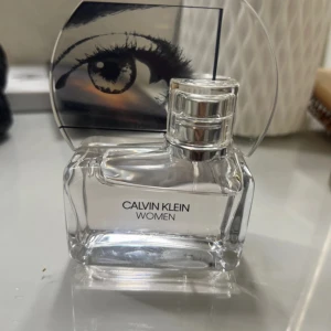 Calvin Klein Women parfym - Stilren parfymflaska från Calvin Klein Women med transparent glas och silvrig kork. Flaskan har en unik design med ett öga på locket, vilket ger en modern och konstnärlig känsla. Perfekt för dig som gillar minimalistisk och trendig stil.