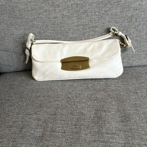 Vit axelväska från Fiorelli - Snygg vit axelväska från Fiorelli med stilren design och justerbart axelband. Väskan har ett elegant metallspänne framtill med märkets logga och är tillverkad i äkta läder .  Perfekt storlek för det viktigaste och passar till många outfits.