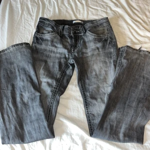 Grå jeans med kontrastsömmar - Säljer ett par grå bootcut-jeans med tydliga vita kontrastsömmar och snygga bakfickor med lock och knapp. Jeansen har klassisk femficksdesign. Materialet är denim och färgen är tvättad grå, vilket ger en cool, avslappnad look. 💓