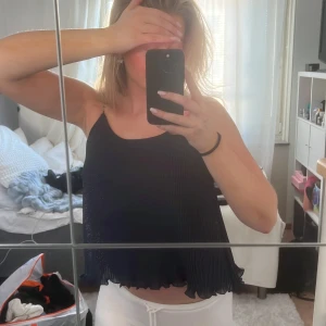 Marinblått glittrig linne - Snygg marinblått glittrig linne med plisserad design och volangdetaljer vid nederkanten. Perfekt för en kväll ute eller en festlig tillställning. Den är ärmlös och har en lös passform som ger en bekväm och stilren look.