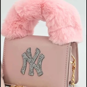 Rosa handväska med pälsdetalj - Snygg rosa handväska med en fluffig pälsdetalj på handtaget och en glittrig logotyp framtill. Väskan har en guldfärgad kedja som axelrem och är perfekt för att ge en lyxig touch till din outfit.