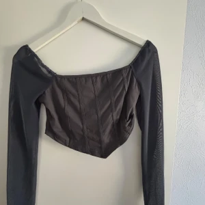 Svart korsettopp med långa ärmar - Snygg grå korsettopp med långa ärmar och offshoulder-design. Toppen har en croppad passform och är perfekt för en trendig look. Den har en stilren och elegant design som passar till många olika tillfällen.