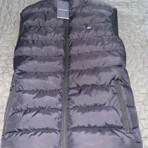 Svart dunväst från Tommy Hilfiger - Snygg svart dunväst från Tommy Hilfiger med dragkedja och diskret logga på bröstet. Perfekt för lager-på-lager under kyliga dagar. Västen har praktiska fickor med dragkedja.