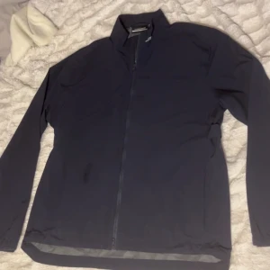 Mörkblå golfjacka från J.Lindeberg - Säljer en stilren mörkblå golfjacka från J.Lindeberg. Jackan har en dragkedja framtill och är tillverkad i polyester, vilket gör den lätt och bekväm.  Snålt använd. 