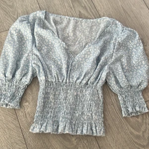 Blå blus med leopardmönster - Säljer en söt blå blus med vitt leopardmönster. Blusen har en v-ringad design och trekvartsärmar. Den smockade midjan ger en fin passform och volangdetaljerna vid ärmslut och nederkant ger en extra touch. Perfekt för en stilren look!