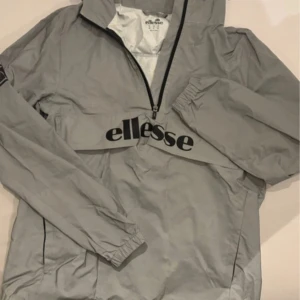 Grå vindjacka från Ellesse - Säljer en grå vindjacka från Ellesse med dragkedja och huva. Jackan har långa ärmar och ett stort tryck med märkesnamnet på framsidan. Perfekt för blåsiga dagar. Säljer den på grund av att den var för stor för mig när jag köpte den från någon annan. Skicket är 8/10. 