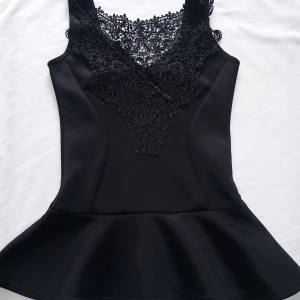 Svart topp med spetsdetaljer från Club L - Elegant svart topp från Club L med vacker spetsdetalj vid halsringningen. Toppen har en figurnära passform och är ärmlös, vilket ger en stilren och sofistikerad look. Perfekt för en kväll ute eller en speciell tillställning.