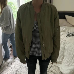 Olivgrön bomberjacka - Säljer en olivgrön bomberjacka med dragkedja och ribbade muddar. Jackan har en klassisk bomberstil med fickor på sidorna och en ärmficka med dragkedja. Insidan är fodrad med orange tyg för en cool kontrast.