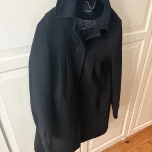 Svart kappa från Twist & Tango - Finns att hämta på Kungsholmen. Elegant svart kappa med en stilren design. Kappan har långa ärmar och en enkel knapp framtill. 