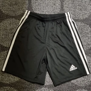 Svarta träningsshorts från Adidas - Snygga svarta träningsshorts från Adidas med klassiska vita ränder längs sidorna. Tillverkade i ett lätt och bekvämt syntetmaterial, perfekt för sport och träning. Adidas-loggan syns på benet och de har en elastisk midja för optimal passform.
