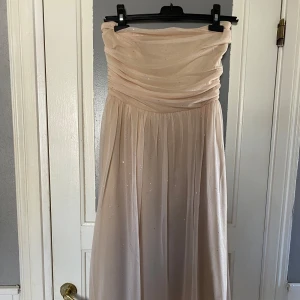 Champagnefärgad axelbandslös balklänning - Elegant axelbandslös klänning i en vacker champagnefärg. Klänningen har en draperad överdel och en lång, flödande kjol med subtila glittriga detaljer. Perfekt för balen.