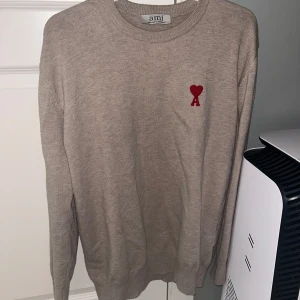 Beige tröja från Ami - Säljer en stilren beige tröja från Ami med ett rött hjärta broderat på bröstet. Tröjan har långa ärmar och en rund halsringning. Perfekt för en avslappnad och trendig look.