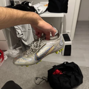 Fotbollsskor från Nike - Säljer ett par fotbollsskor från Nike med en futuristisk design i silver och regnbågsfärger. Skorna har snörning och rund tå, samt en sula med gula dobbar för bra grepp på planen. Köpta för 2799 men är ganska använda och lite skrapsår på ena sidan men är skit bra att köra med fortfarande!