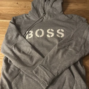 Grå hoodie från Hugo Boss - Säljer en stilren grå hoodie från Hugo Boss med stor vit text 'BOSS' på bröstet. Tröjan har en klassisk design med känguruficka och justerbar huva med dragsko. Perfekt för en avslappnad look.