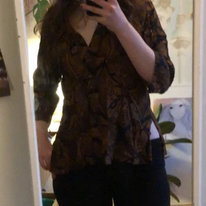 Mönstrad blus från Manuheali'i - Säljer en unik mönstrad blus från Manuheali'i i bruna och svarta toner. Blusen har en v-ringad design med trekvartsärmar och en lätt volangdetalj. Perfekt för att ge en bohemisk touch till din outfit.