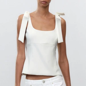 Vit ärmlös topp med rosetter från Zara - Snygg vit ärmlös topp med breda axelband och dekorativa rosetter. Toppen har en stilren design med en fyrkantig urringning och en figurnära passform. Perfekt för en elegant och modern look. Helt ny 