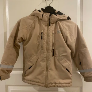 Beige jacka med huva - Säljer en beige jacka med huva och dragkedja. Jackan har långa ärmar med reflexdetaljer och flera praktiska fickor med dragkedjor. Perfekt för kyligare dagar.