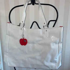 Vit axelväska från Nina Ricci - Snygg vit axelväska från Nina Ricci med ett rött äpple som dekoration. Väskan har en rymlig design med röda detaljer inuti och en praktisk dragkedja. Mått 48 *45 cm.Perfekt för att bära med sig allt man behöver under dagen.