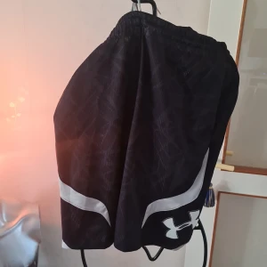 Svarta shorts från Under Armour - Säljer ett par svarta shorts från Under Armour med vitt mönster och logga. De har en loose passform och är perfekta för träning. Shortsen har elastisk midja för extra komfort.