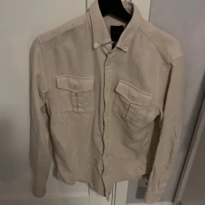 Beige skjorta från Oscar Jacobson - Säljer en stilren beige skjorta från Oscar Jacobson i storlek M. Skjortan har långa ärmar och två bröstfickor med knappar. Perfekt för en elegant look.