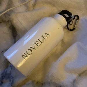 Vit vattenflaska från Novelia - Snygg och praktisk vit vattenflaska från Novelia. Flaskan har en svart skruvkork med karbinhake, perfekt för att fästa på väskan eller bältet. Stilren design med Novelia-logga i svart text. Kan även användas för kaffet på morgonen 😅