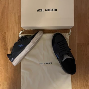 Axel Arigato skor - Snygga marinblå sneakers från Axel Arigato med en blå fågelbrodering på sidan. Skorna har en vit sula och snörning framtill. Perfekta för en stilren look.