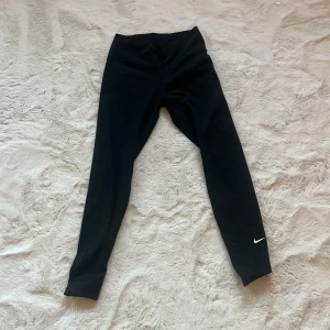 Svarta leggings från Nike - Säljer ett par svarta leggings från Nike med Dri-FIT-teknologi. De har en hög midja och en liten Nike-logga på benet. Perfekta för träning och bekväma att bära.