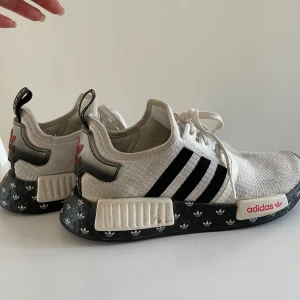 Adidas NMD R1 sneakers - Snygga Adidas NMD R1 sneakers i vitt med svarta och röda detaljer. Skorna har en unik svart sula med vita mönster och klassiska tre ränder på sidan. Perfekta för en stilren look.