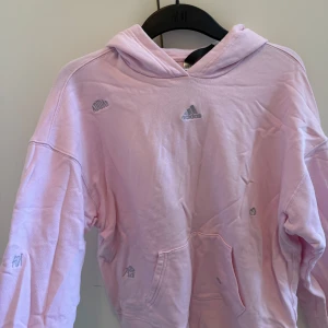 Rosa hoodie från Adidas - Säljer en snygg rosa hoodie från Adidas med logotypdetaljer. Den har en klassisk känguruficka och en bekväm huva. Perfekt för en avslappnad stil.