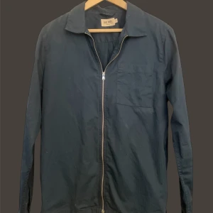 East West Overshirt - Snygg mörkblå overshirt med dragkedja framtill. Den har en klassisk krage och en bröstficka. Perfekt för lager-på-lager-stil.