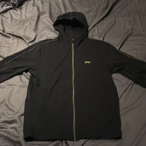 Svart boss hoodie med zip - Säljer en svart hoodie med dragkedja och huva från Boss med färgtemat svart och guld