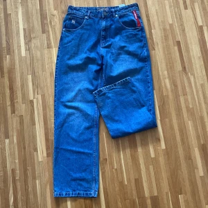 Blå baggy jeans från DNGRS - Säljer ett par blå baggy jeans från DNGRS med snyggt broderad bakficka och röd detalj vid fickan fram. Jeansen har en klassisk femficksdesign och knappgylf. Perfekta för en avslappnad stil.