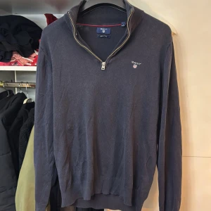 Mörkblå tröja med dragkedja från GANT - Säljer en stilren mörkblå tröja från GANT med dragkedja vid halsen. Tröjan är tillverkad i en blandning av bomull och ull, vilket gör den både bekväm och varm. Perfekt för kyligare dagar.