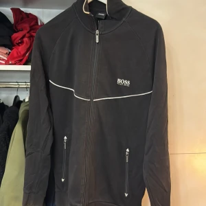 Hugo Boss tröja  - Snygg svart jacka från Boss med dragkedja och vita detaljer. Jackan har en hög krage och två fickor med dragkedjor framtill. Perfekt för en stilren look.