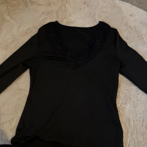Svart långärmad topp med spetsdetaljer - Snygg svart långärmad topp med v-ringad spetsdetalj. Perfekt för en elegant look. Passar bra till både jeans och kjol.