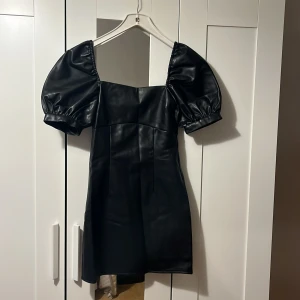 Fest klänning - Säljer en svart kort klänning med markerade puffärmar och fyrkantig urringning. Klänningen är gjord i skinnimitation och har snörningsdetaljer i ryggen. Helt ny!