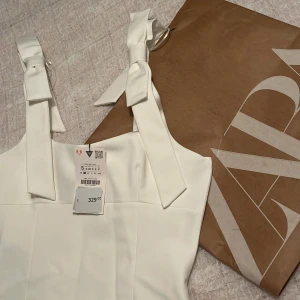 Vit linnetopp med rosettband från Zara - Säljer en stilren vit linnetopp från Zara med breda axelband som knyts i rosetter. Toppen har en rak passform och är perfekt för varma dagar. Snygg och enkel att matcha till olika outfits. Den är inte använd eftersom att vi köpte fel storlek.