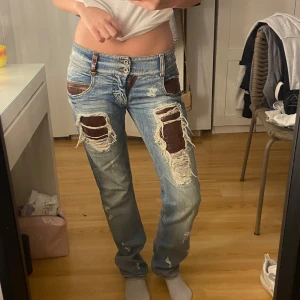 Ljusblå lågmidjade jeans - Säljer ett par ljusblå jeans med bruna detaljer och stora slitningar framtill. Jeansen har raka ben och bruna lappar vid fickor och lår. Snygga kontrastsömmar och cool vintagekänsla.