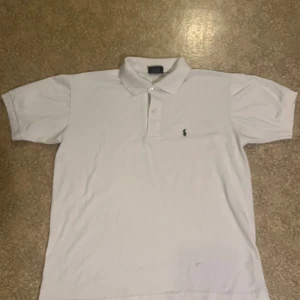 Vit pikétröja Polo Sport - Klassisk vit pikétröja från Polo Sport med krage och knappar framtill. Passar även M. Tröjan har en liten broderad logga i mörkblått på bröstet och är tillverkad i 100% bomull. Perfekt för en stilren och sportig look.