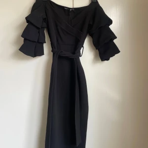 Omlottklänning med volangärmar - Omlottklänning med trekvartsärm i flera volanger. Klänningen har omlottliv och ett matchande bälte i midjan. Från booho i storlek 36.