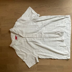 Vit linneskjorta från RYVLS - Säljer en luftig och stilren vit skjorta från RYVLS i linne. Skjortan har korta ärmar, klassisk krage och knäppning framtill med vita knappar. Perfekt för varma dagar och enkel att matcha med olika outfits.