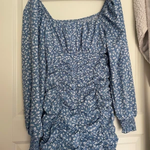 Blå blommig klänning med rynkade detaljer - Supersöt blå klänning med vita små blommor och rynkade detaljer framtill. Klänningen har långa ärmar med resår vid ärmslut och en fyrkantig ringning. Perfekt för vår och sommar!