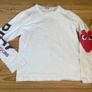 Vit långärmad tröja från Comme des Garçons Play - Vit långärmad tröja från Comme des Garçons Play med svart text på ena ärmen och det klassiska röda hjärtat med ögon på andra ärmen. Storlek 158-164 vilket motsvarar L i barnstorlek. Rund halsringning och avslappnad passform. Perfekt för dig som gillar streetwear och unika detaljer.