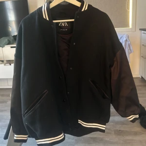 Svart varsityjacka från Zara - Säljer en svart varsityjacka från Zara med vita ränder på krage, ärmslut och nederkant. Jackan har långa ärmar i kontrasterande material och två snedställda fickor framtill. Klassisk college-look med knappar framtill. Strl xs men är over sides