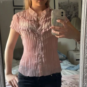 Ljusrosa blus med spets och volang - Säljer en ljusrosa blus med korta ärmar och knappar framtill. Blusen har fina spetsdetaljer och volanger upptill, samt ett lätt skrynklat tyg. 