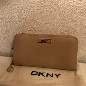 Beige plånbok från DKNY - Snygg beige plånbok från DKNY med dragkedja runtom och guldig logga framtill. Insidan är fodrad i stark rosa färg med flera fack för kort och mynt. Perfekt accessoar för att hålla ordning på dina värdesaker.