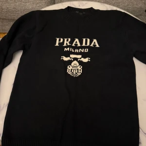 Svart stickad tröja från Prad*  - Säljer en svart stickad tröja från Prad*  med vit text 'PRAD* MILAN*' och emblem på bröstet. Tröjan har rund halsringning och långa ärmar. Perfekt för dig som gillar klassiska designerplagg. Bra skick. Skickar alltid inom 24 timmar. 