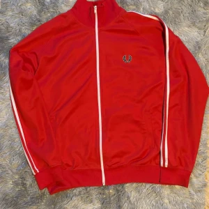 Röd track jacket från Fred Perry - Röd track jacket från Fred Perry med vita ränder längs ärmarna och vit dragkedja. Jackan har ståkrage, ribbade muddar och Fred Perry-logga broderad på bröstet. Perfekt för en sportig och avslappnad stil.