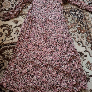 Blommig långklänning med volang - Säljer en långklänning med blommigt mönster i rosa, svart och vitt. Klänningen har långa ärmar och volangdetaljer nertill. Perfekt för dig som gillar romantisk och bohemisk stil.