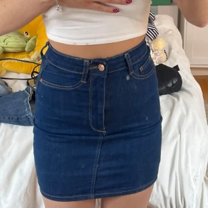 Mörkblå jeanskjol från Gina Tricot - Säljer en klassisk mörkblå jeanskjol från Gina Tricot. Kjolen har synliga sömmar och knapp- samt dragkedjestängning framtill. Snygg och enkel modell som är lätt att matcha till olika toppar. Säljer då den knappt har använts och e liten tight. 